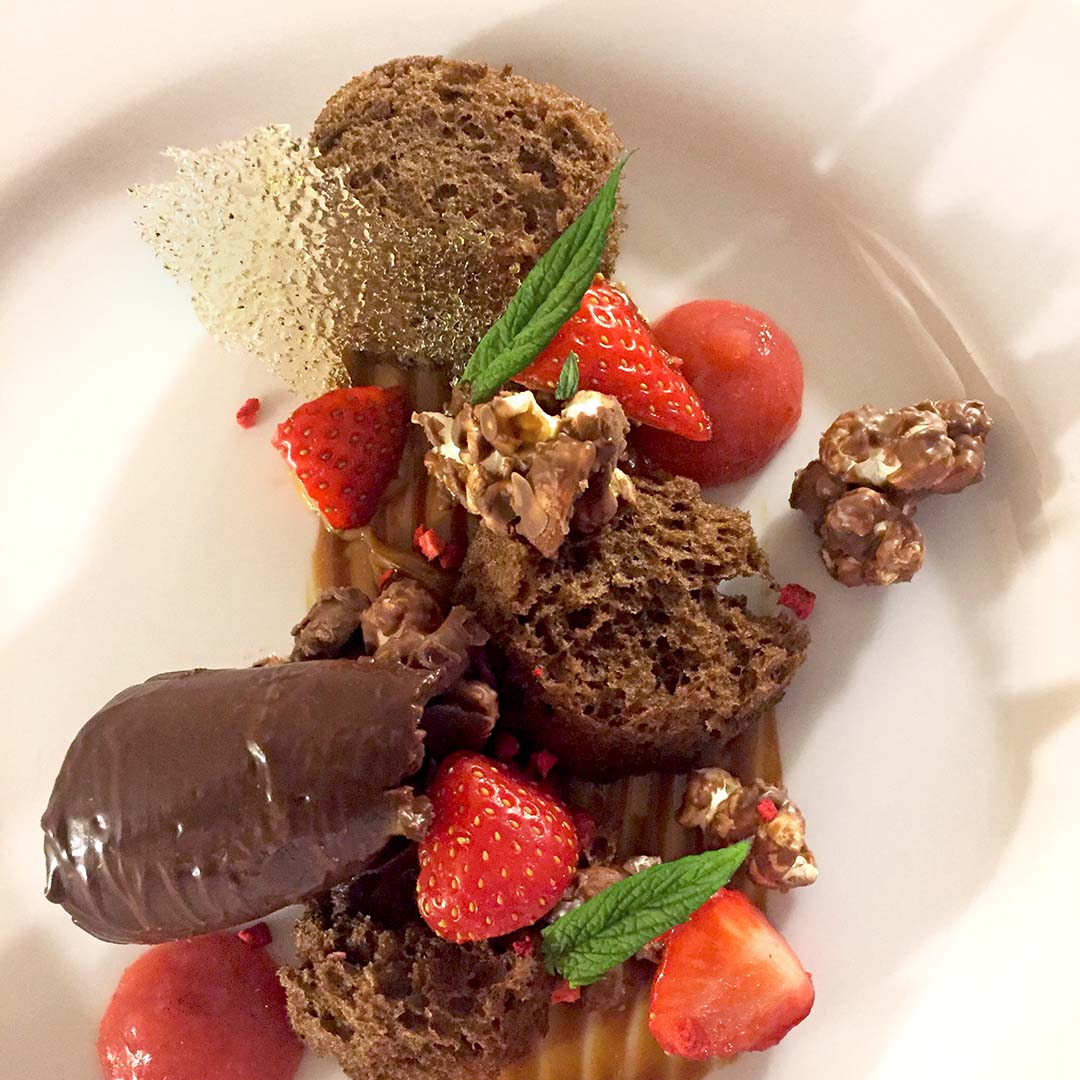 A plated dessert featuring chocolate sponge, chocolate mousse, fresh strawberries, candied nuts, a sugar shard, caramel sauce, and a mint sprig, all artfully arranged on a white plate.