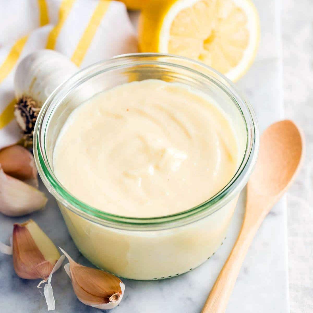 A small glass jar filled with creamy aioli sits on a marble surface, surrounded by garlic cloves, a wooden spoon, a halved lemon, and a white cloth with yellow stripes.