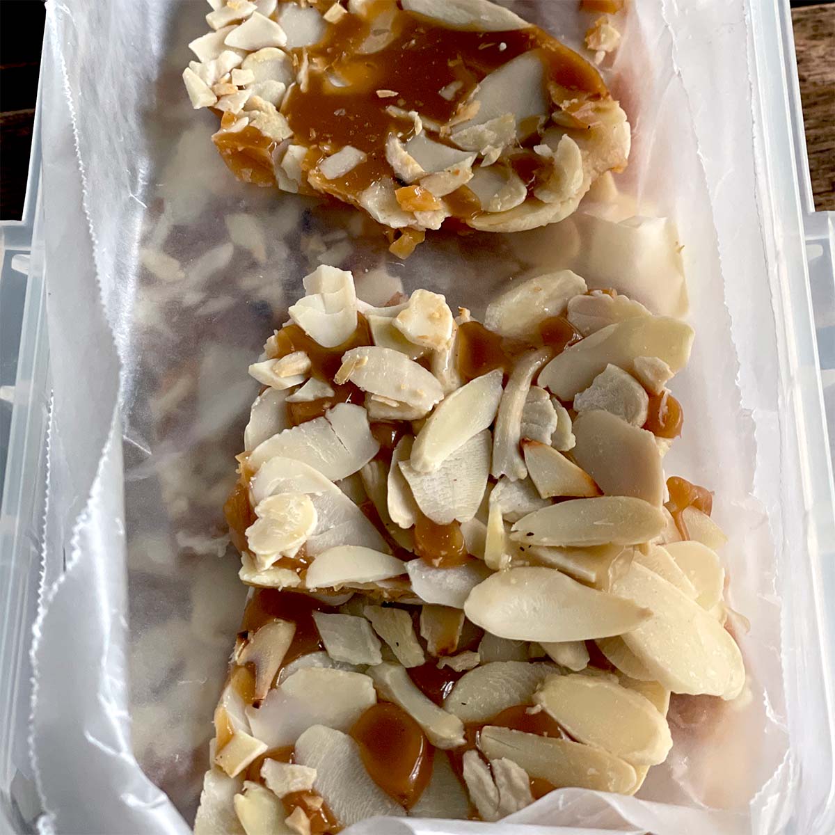 Two pieces of almond brittle with caramel and sliced almonds are placed on greaseproof paper inside a rectangular container.