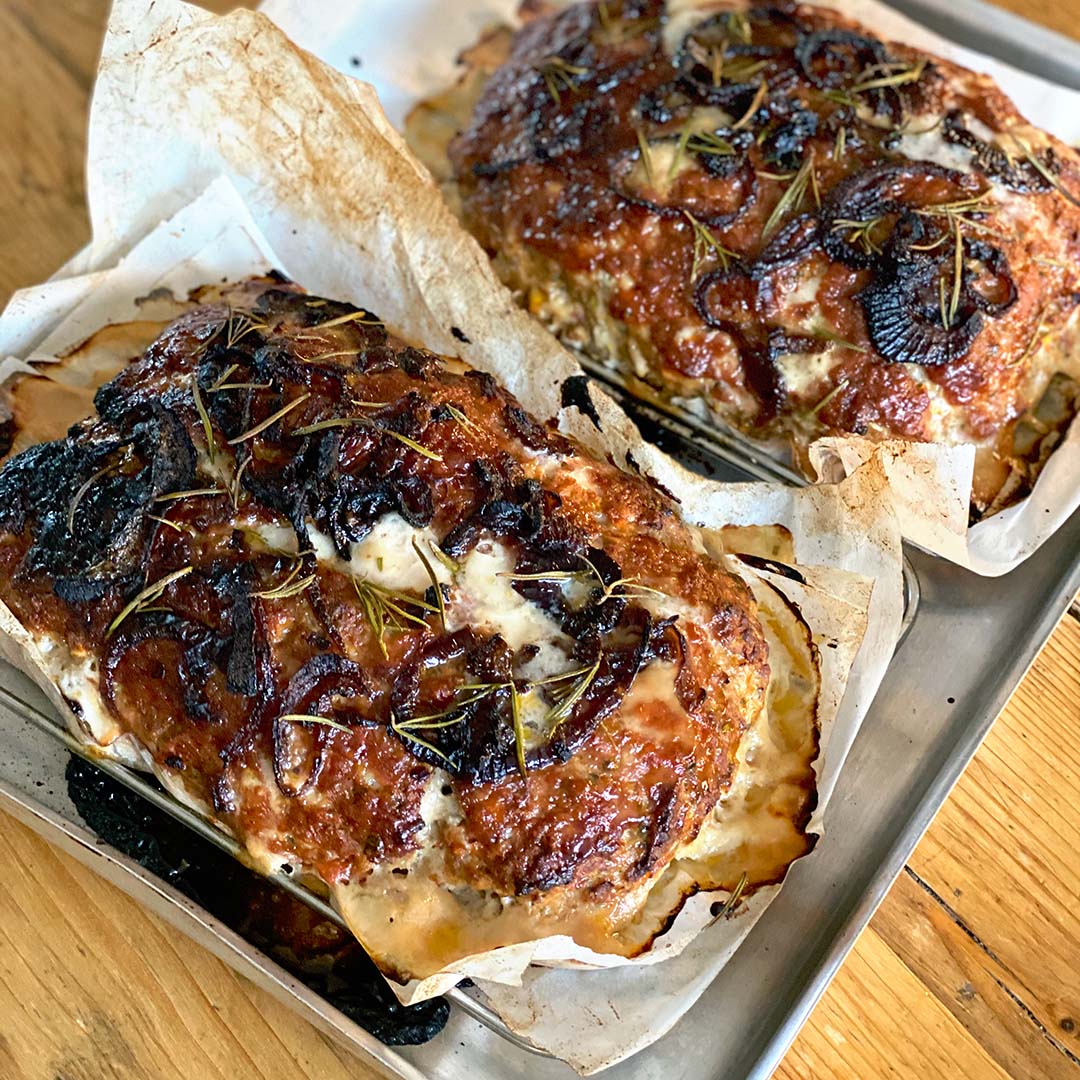 Two baked meatloaves with caramelised, browned tops, garnished with herbs and sliced onions, resting on parchment-lined baking trays. The meatloaves are golden with visible seasoning and a rustic appearance.