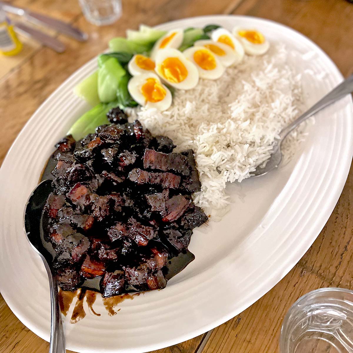 A white oval platter with steamed white rice, soy-braised pork belly in dark sauce, sliced soft-boiled eggs, and blanched green vegetables, served with a large serving spoon.