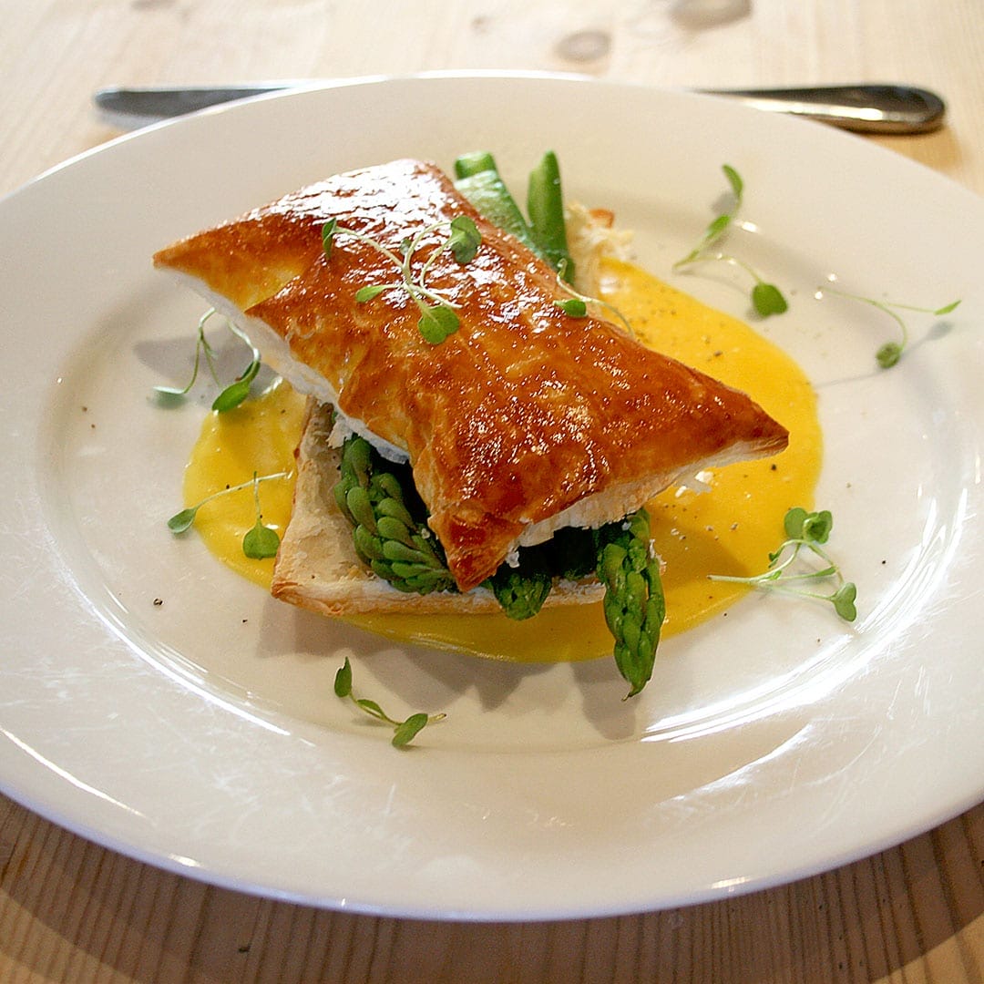 A golden puff pastry sits atop fresh asparagus spears and sauce on a white plate, garnished with microgreens, on a light wooden table. A fork and knife are in the background.