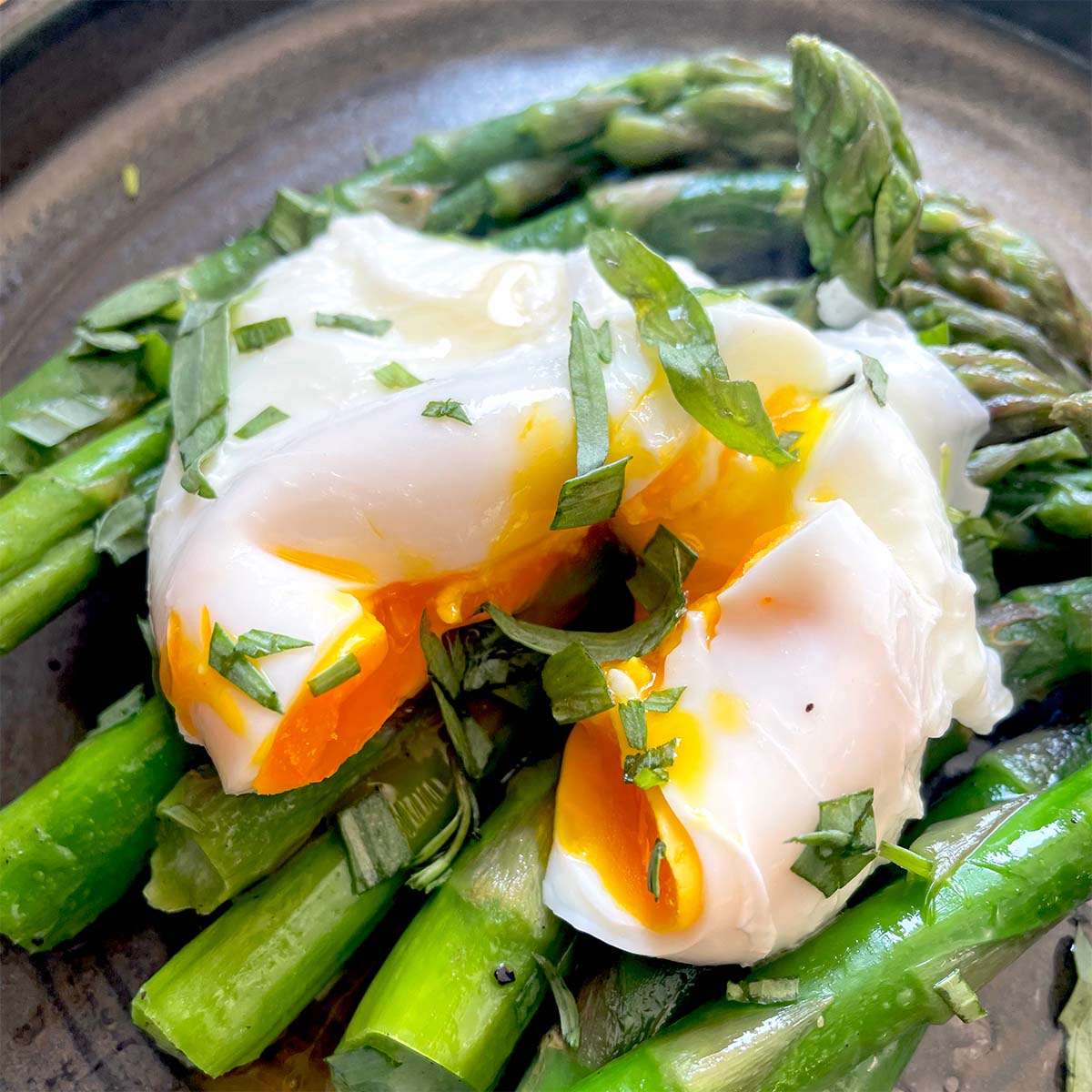 A poached egg with a runny yolk sits atop bright green asparagus spears, garnished with chopped fresh herbs, on a dark plate.