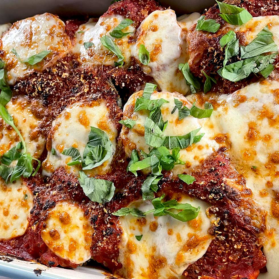 Close-up of baked chicken parmigiana topped with melted mozzarella cheese, tomato sauce, grated parmesan, and fresh basil leaves. The dish has a golden, bubbly, and slightly browned appearance.