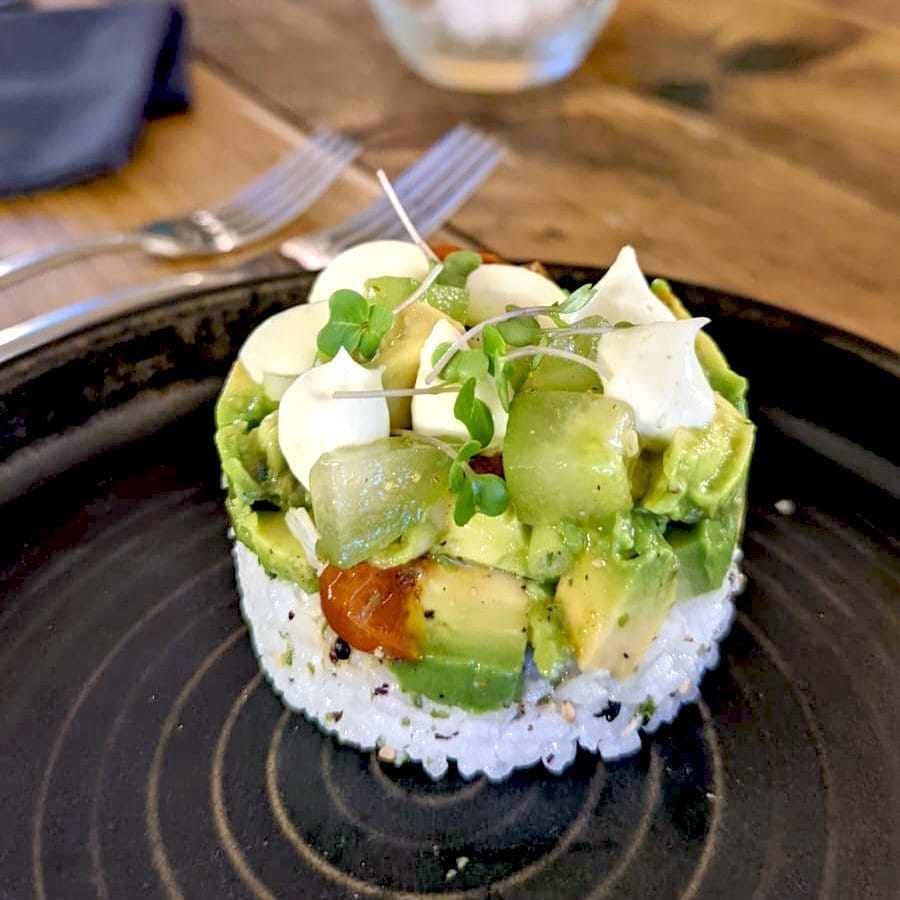 A round stack of sushi rice topped with diced avocado, kiwi, cherry tomatoes, microgreens, and a drizzle of creamy sauce, served on a black plate with a fork and knife in the background.