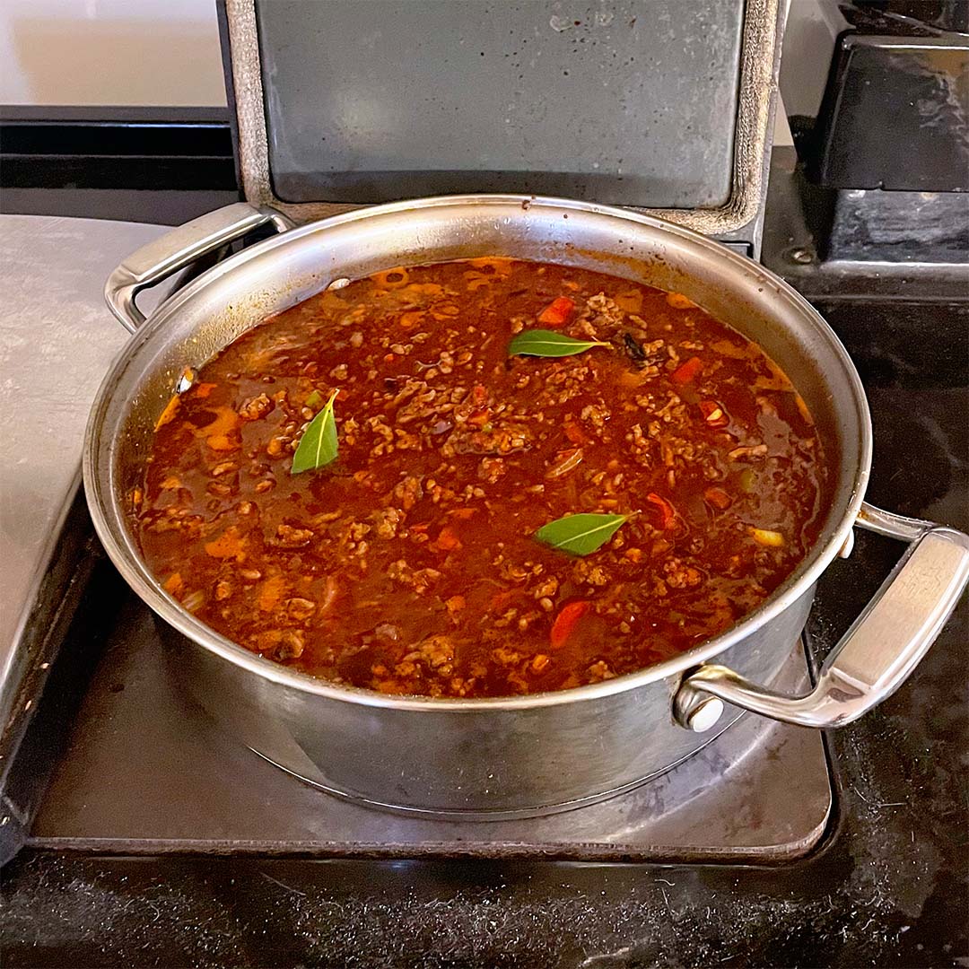 A large stainless steel saucepan on a hob contains simmering meat sauce with visible minced beef, tomato pieces, and bay leaves floating on the surface.