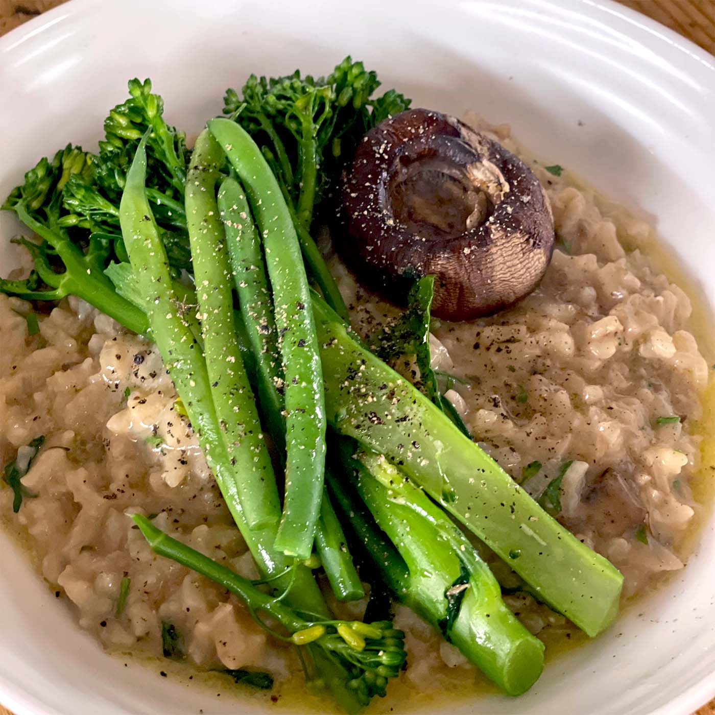 A bowl of creamy risotto topped with steamed tenderstem broccoli and a roasted mushroom, garnished with cracked black pepper and fresh herbs.
