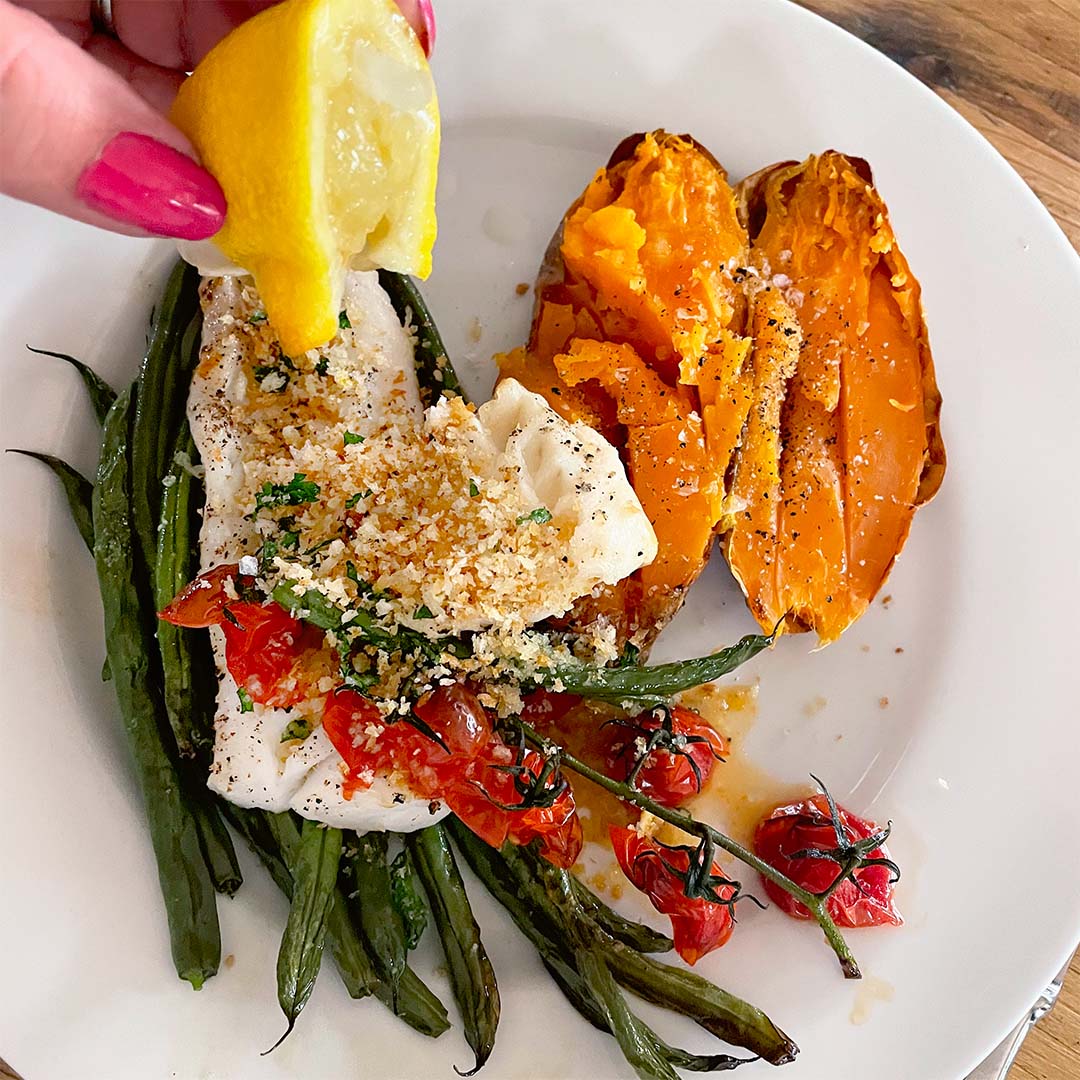 A hand squeezes a lemon over a plate with baked fish topped with breadcrumbs and cherry tomatoes, roasted green beans, and a split baked sweet potato sprinkled with seasoning.