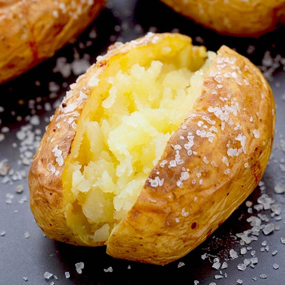 A baked potato split open to reveal its fluffy interior, with coarse salt sprinkled on its crispy golden skin and on the surrounding dark surface.