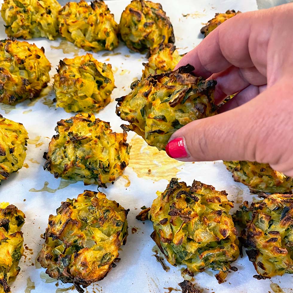 A hand with pink nail varnish picks up a golden-brown baked vegetable fritter from a tray lined with baking paper, surrounded by several similar fritters.