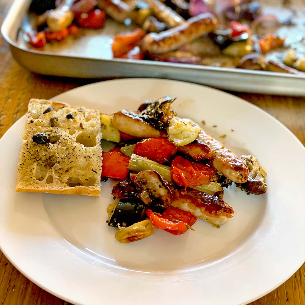 A white plate with roasted vegetables, sausage, and a slice of olive focaccia bread. A baking tray with more roasted vegetables and sausages is in the background on a wooden table.