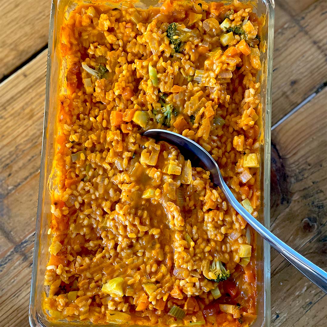 A glass dish filled with creamy orange risotto containing visible pieces of vegetables, such as carrots and broccoli, with a silver spoon resting inside. The dish is placed on a wooden surface.