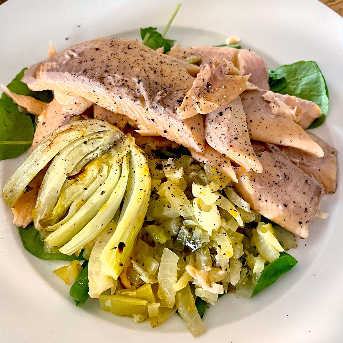 A plate of poached fish fillets topped with black pepper, served on a bed of sautéed greens, onions, and a wedge of cooked artichoke. The dish is arranged on a white plate.