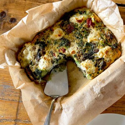 A baked vegetable and cheese frittata in a parchment-lined dish, with a serving spatula and a wedge already removed, sits on a wooden table.