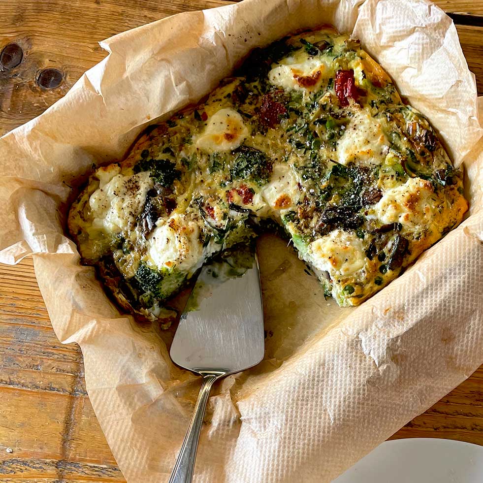 A baked vegetable and cheese frittata in a parchment-lined dish, with a serving spatula and a wedge already removed, sits on a wooden table.