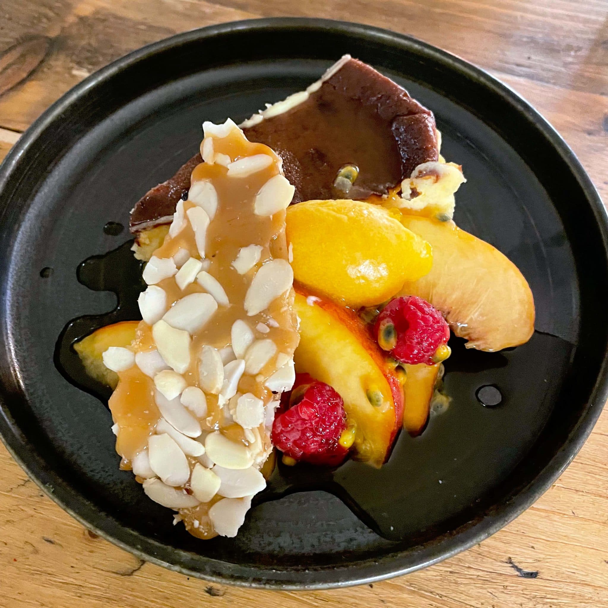 A black plate with a slice of cheesecake, topped with caramel almond brittle, fresh peach slices, raspberries, passion fruit, and drizzled with a dark sauce. The dish is served on a wooden table.