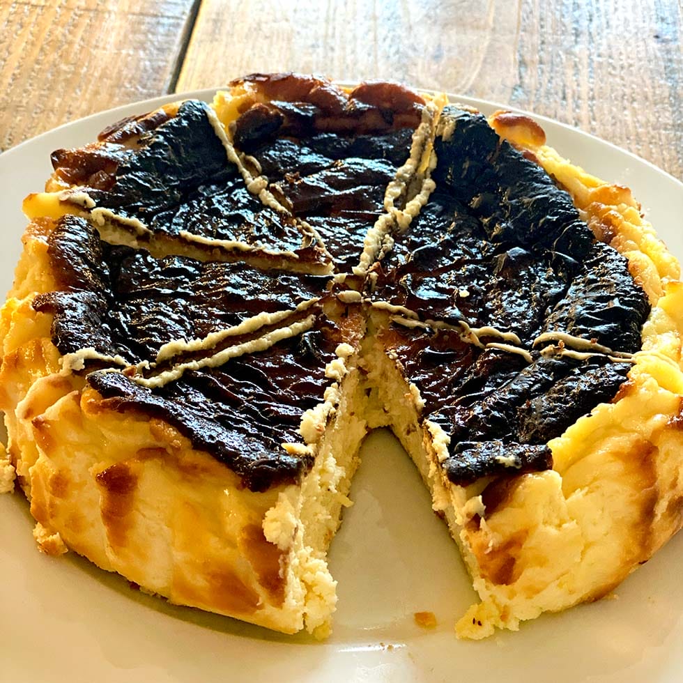A round, rustic cheesecake with a deeply browned, cracked top sits on a white plate. One large wedge is missing, revealing the creamy interior. The dessert is on a wooden surface.