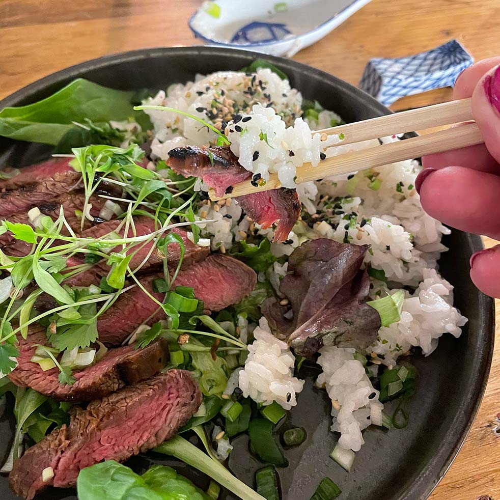 A hand uses chopsticks to pick up a bite of sliced steak and rice from a bowl filled with white rice, sesame seeds, sliced steak, fresh greens, and microgreens.
