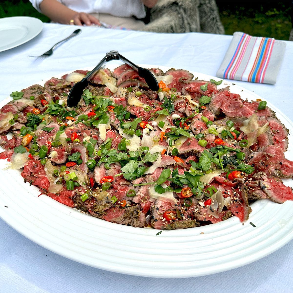 A large white platter of thinly sliced beef carpaccio topped with herbs, red chillies, sesame seeds, and shaved parmesan, with tongs resting on top; a white tablecloth and colourful serviette in the background.