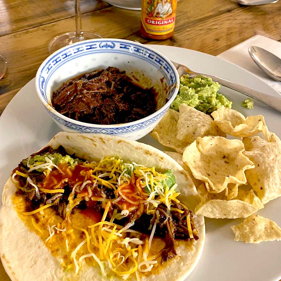 A plate with a soft taco filled with shredded beef, cheese, and sauce, alongside tortilla chips, guacamole, and a bowl of shredded beef. A hot sauce bottle is in the background.