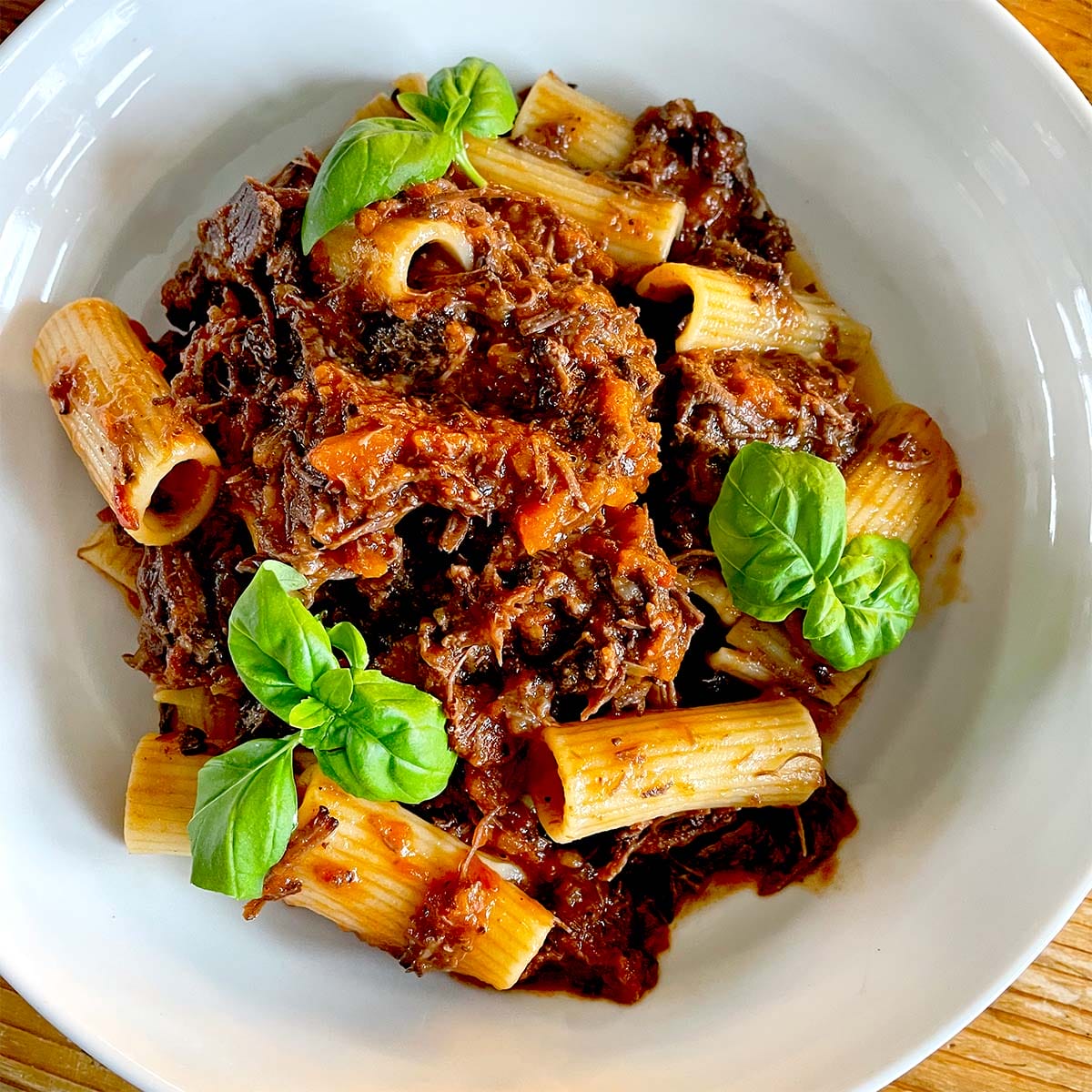 A white bowl filled with rigatoni pasta topped with a rich, chunky meat sauce, garnished with fresh basil leaves.