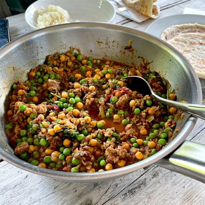 A metal pan filled with a colourful dish of minced meat, green peas, and chickpeas in a tomato-based sauce sits on a wooden table, with a spoon inside. Plates of rice and flatbread are visible in the background.