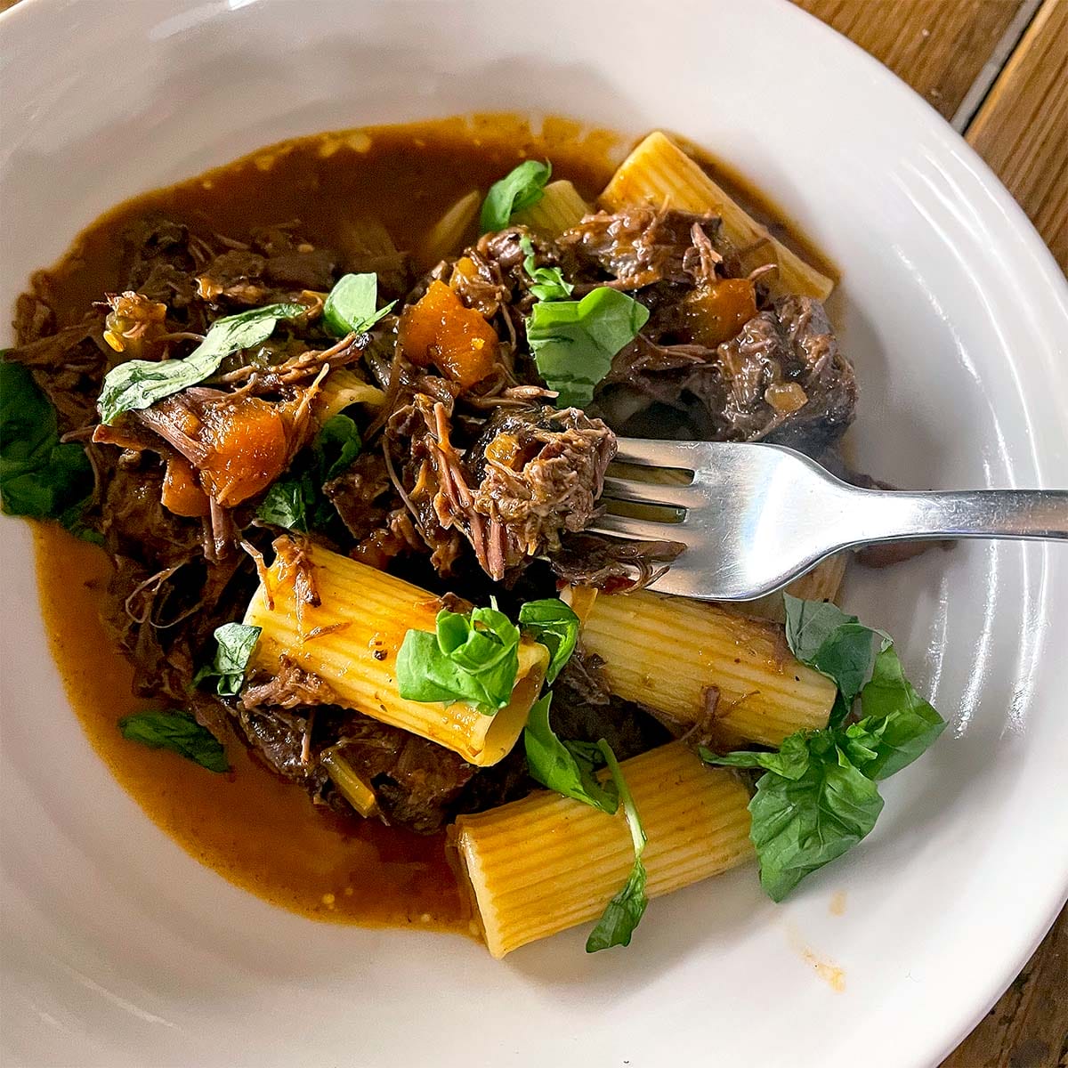 A white plate with rigatoni pasta topped with shredded beef in a rich sauce, diced vegetables, and fresh basil leaves. A fork rests on the plate.