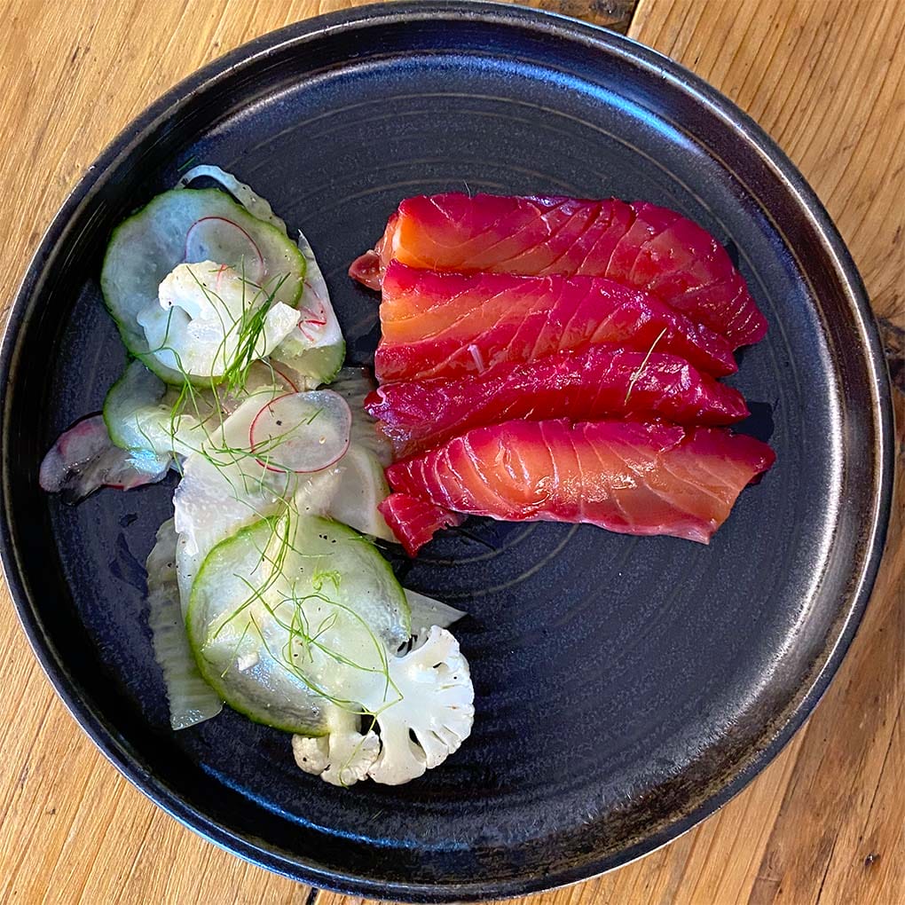 A black plate with slices of beet-cured salmon and a side salad of thinly sliced cucumber, radish, cauliflower, and fresh dill, set on a wooden table.