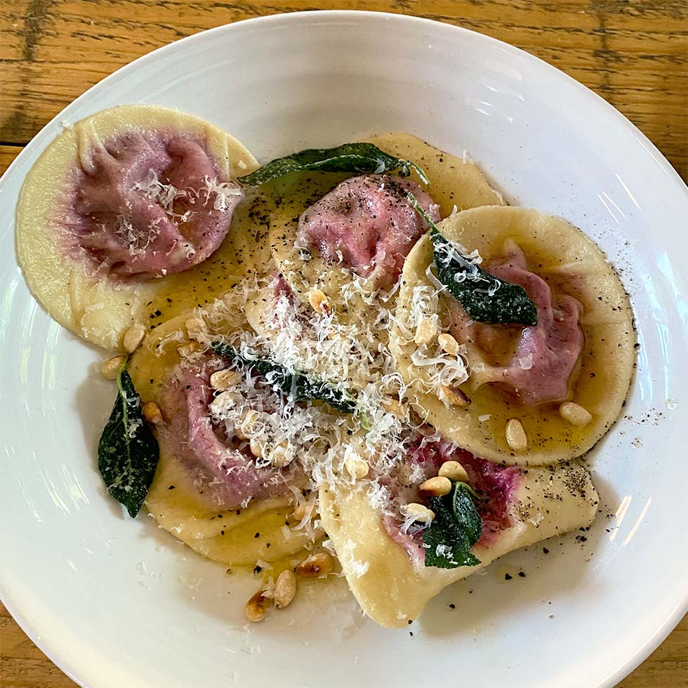 A white plate of round ravioli with purple filling, topped with grated cheese, crispy sage leaves, toasted pine nuts, and black pepper, on a wooden table.