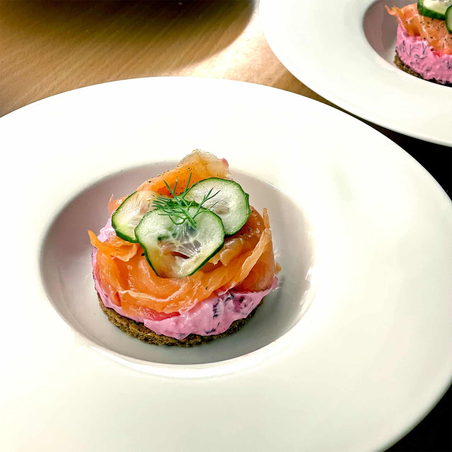 An elegantly plated starter featuring a round base of dark bread topped with a pink creamy spread, slices of smoked salmon, thin cucumber slices, and a sprig of dill, served on a white plate.