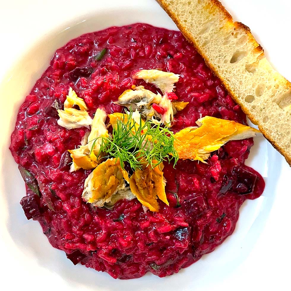 A white bowl of vibrant beetroot risotto, topped with chunks of smoked fish and fresh dill, served with a slice of toasted bread on the side.
