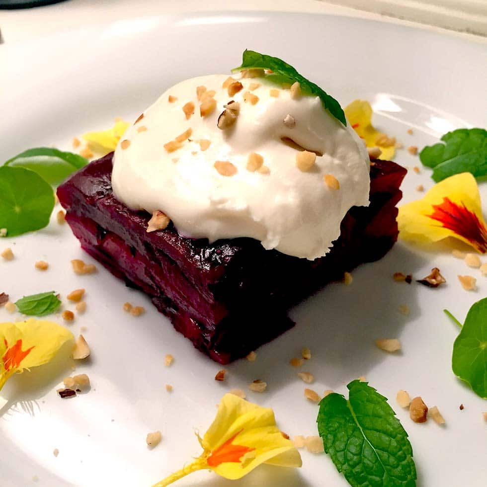 A square slice of beetroot terrine topped with a dollop of cream and chopped nuts, garnished with yellow edible flowers and fresh mint leaves on a white plate.