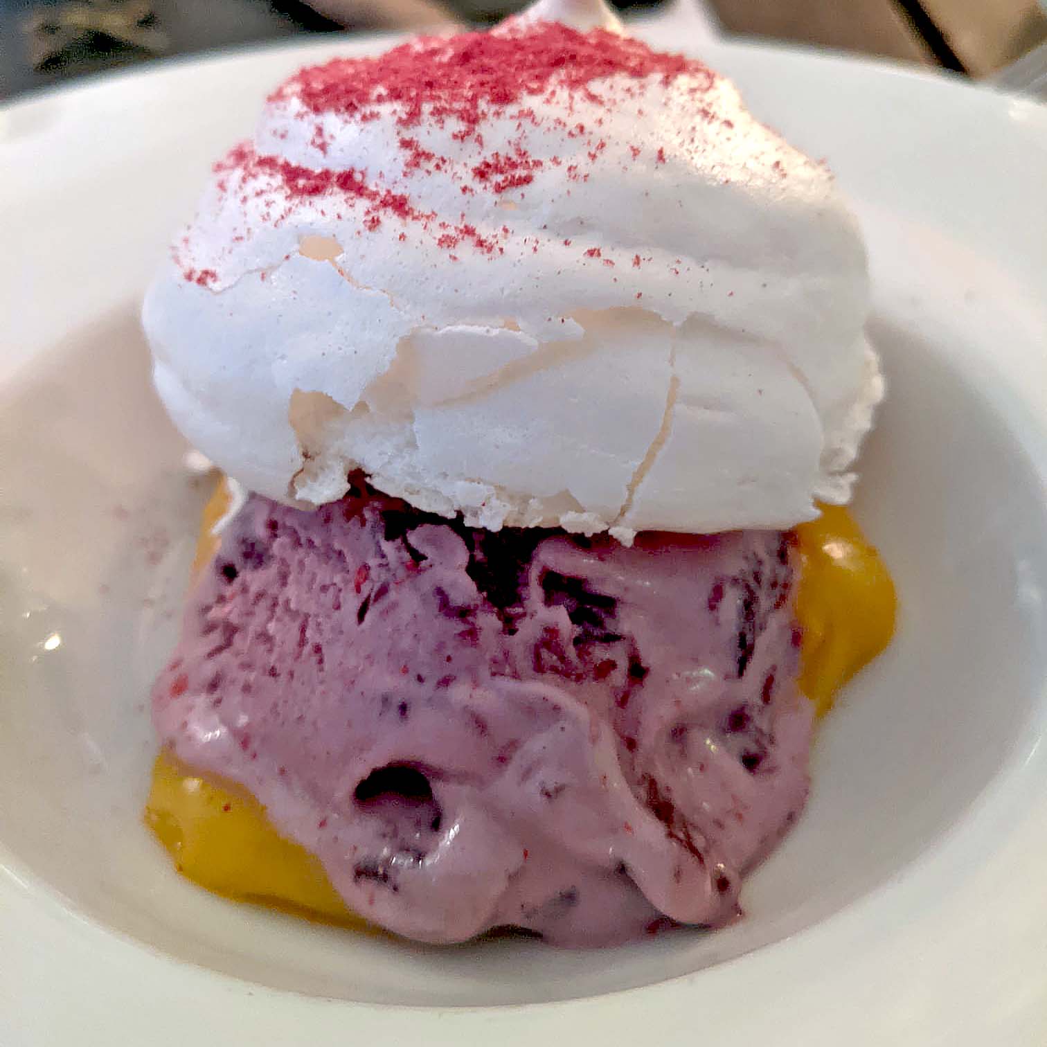 A dessert featuring a scoop of purple berry ice cream on a yellow custard base, topped with a round, crisp white meringue and a sprinkle of pink powder, served in a white bowl.