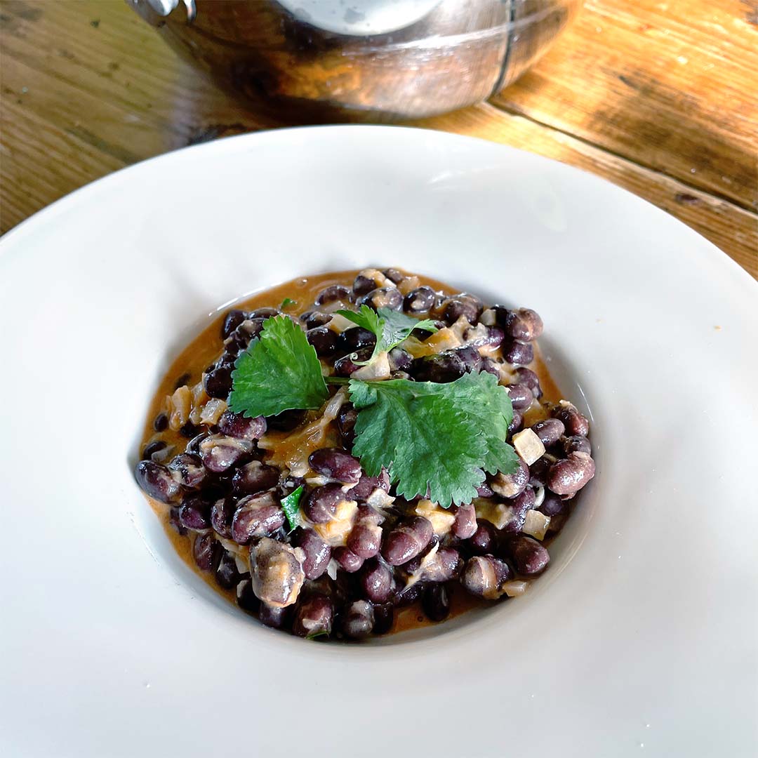 A white bowl filled with creamy black beans garnished with fresh coriander leaves, placed on a wooden table.