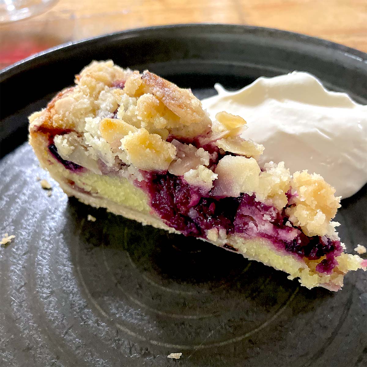 A slice of berry crumble pie with a crumbly topping sits on a dark plate next to a dollop of whipped cream.