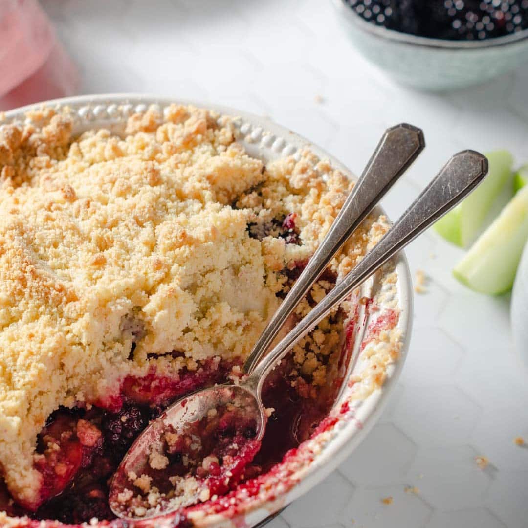A round baking dish filled with berry crumble, topped with a golden, crumbly crust. Two spoons rest inside, and some fruit filling is visible. In the background, a bowl of berries and apple slices are partially seen.
