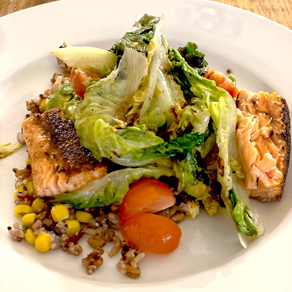 A plate of grilled salmon served with mixed grains, sweetcorn, cherry tomatoes, and sautéed greens, arranged on a white dish.