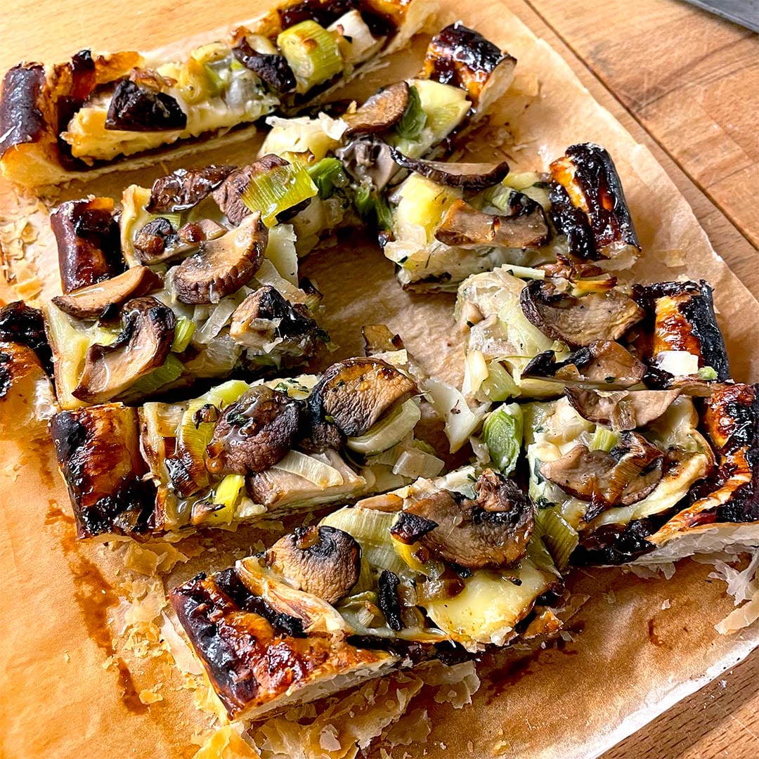 Rectangular slices of savoury tart topped with mushrooms, leeks, and cheese on a sheet of baking parchment, resting on a wooden surface. The tart has a golden, flaky pastry and appears freshly baked.