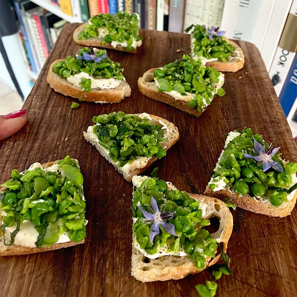 Sliced bread topped with creamy cheese, chopped green peas, herbs, and purple edible flowers arranged on a wooden board, with books visible in the background.