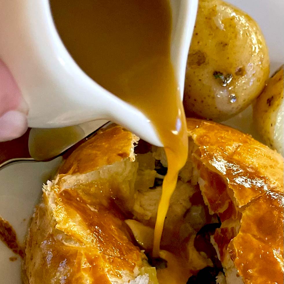 Golden brown gravy is being poured from a small jug over a flaky pastry dish, with roast potatoes visible in the background. The pastry is cracked open, revealing its filling inside.