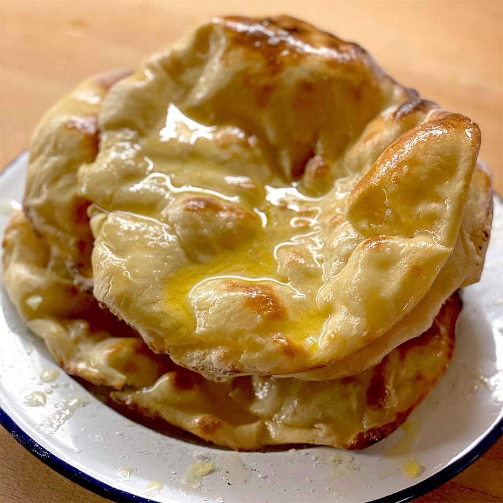A stack of golden, puffy naan breads glistening with melted butter sits on a white plate with a blue rim, resting on a wooden surface.