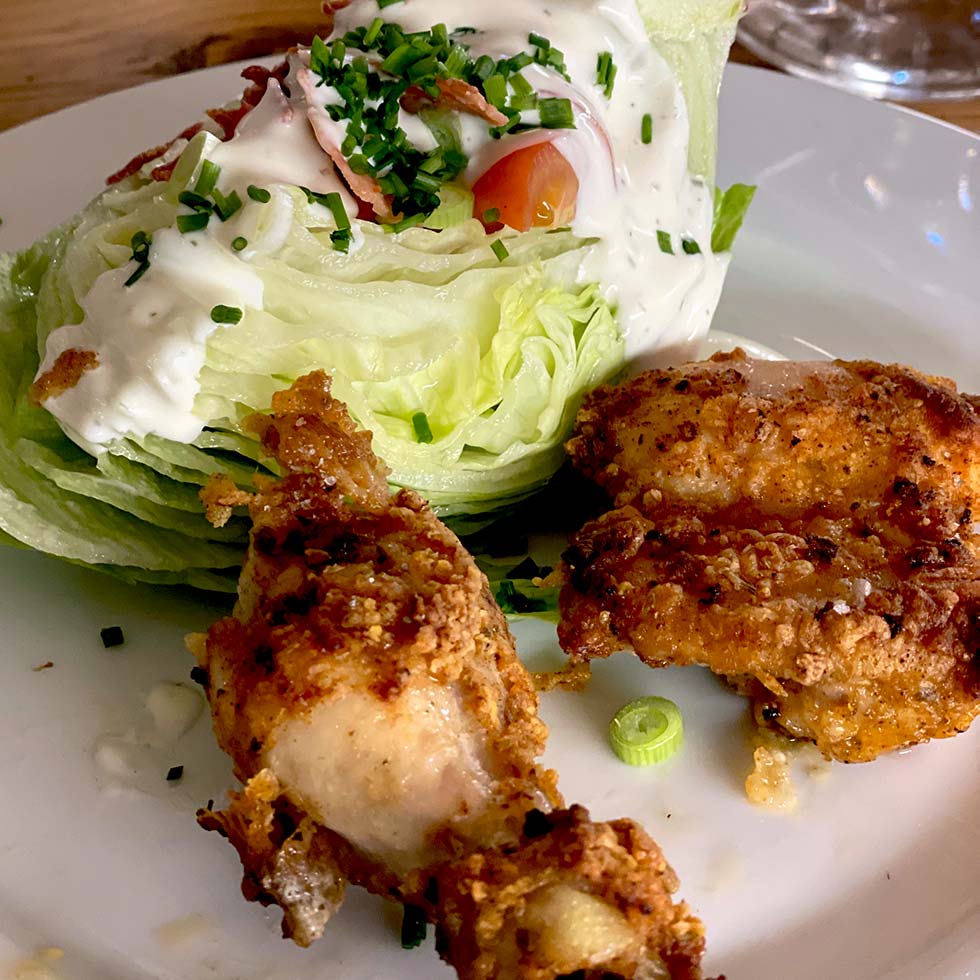 A plate with a wedge of iceberg lettuce topped with creamy dressing, chopped chives, and tomato, served alongside two pieces of crispy fried chicken.
