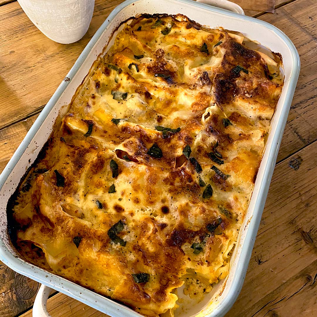 A baked dish of golden-brown lasagne with melted cheese and herbs sits in a white rectangular baking dish on a wooden table.