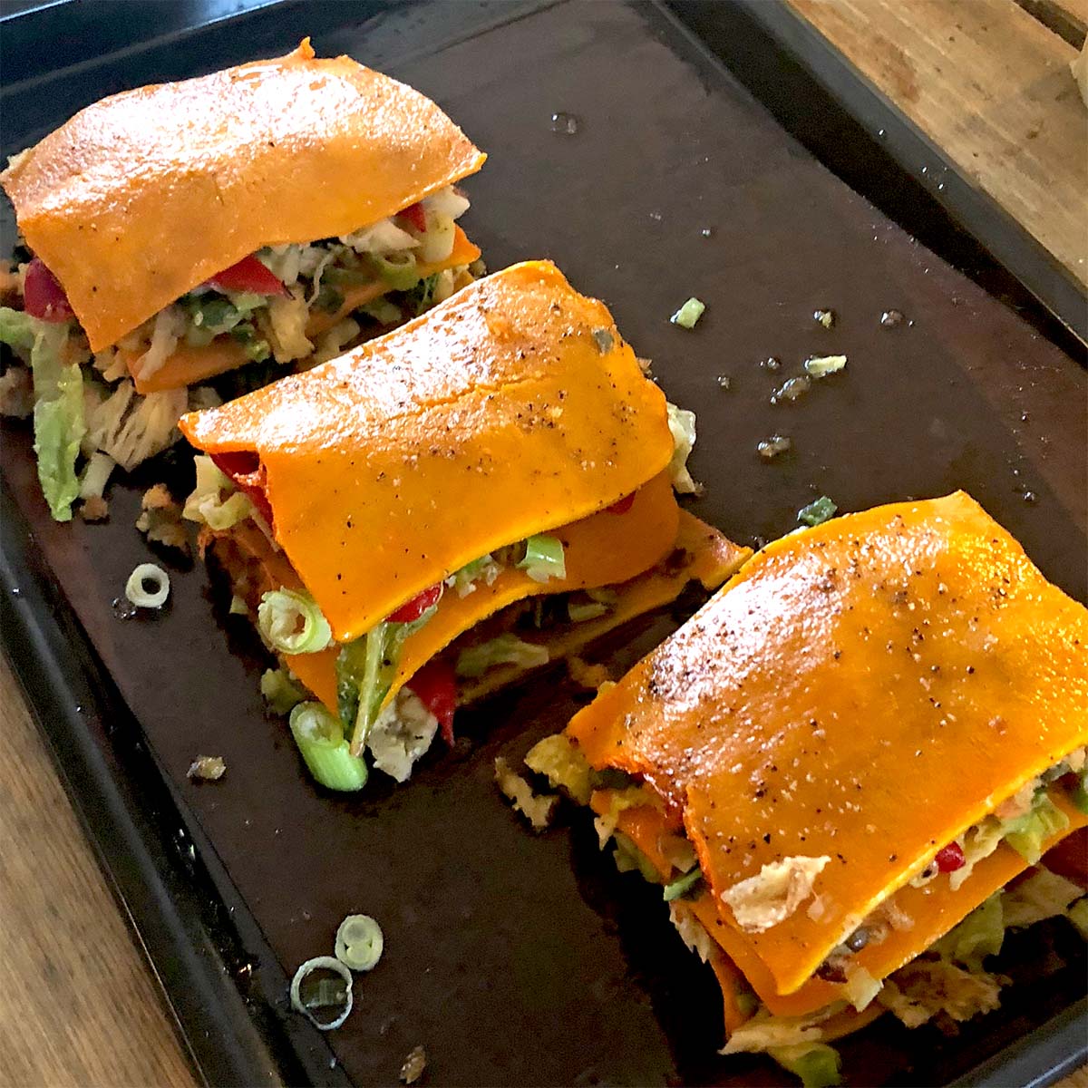 Three servings of vegetable lasagne made with orange squash slices instead of pasta sheets, layered with various vegetables, placed on a dark baking tray.