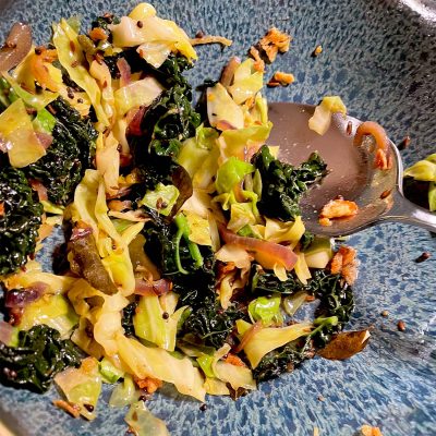 Sautéed cabbage and kale with onions and garlic in a blue speckled bowl, partially eaten, with a metal spoon resting on the side.