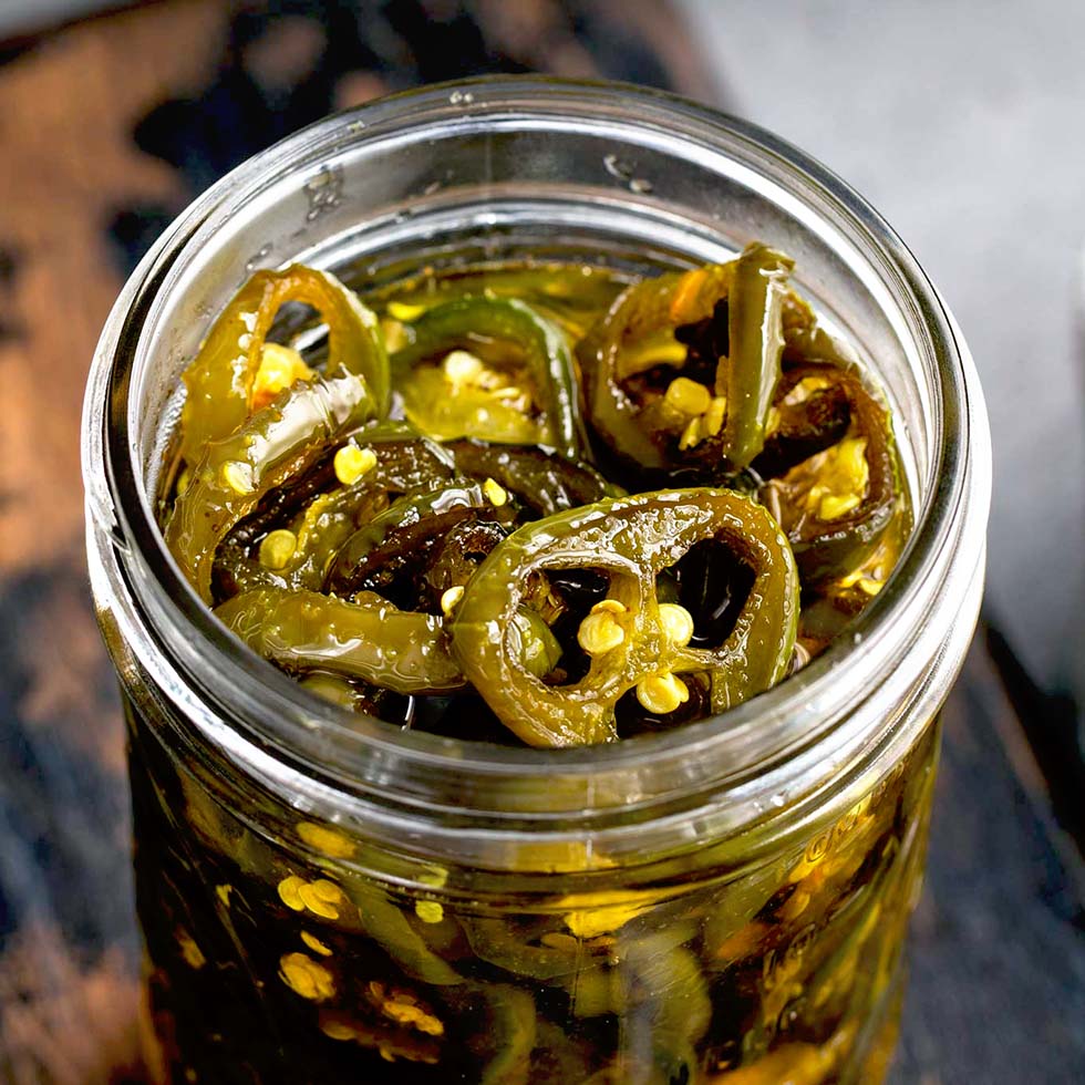 A close-up of a glass jar filled with sliced pickled jalapeños in brine, showing seeds and pepper rings. The jar sits on a dark wooden surface.