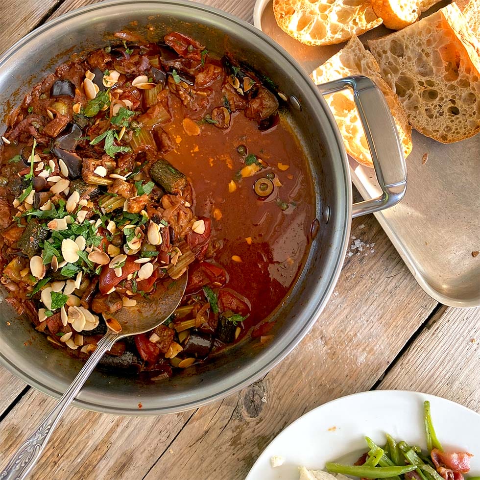 A pot of vegetable stew topped with fresh herbs and sliced almonds sits on a wooden table next to a tray of sliced bread and a plate with green beans.