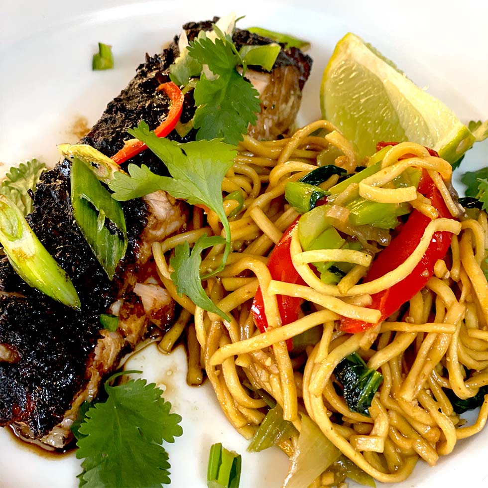 Grilled fish fillet topped with herbs, served alongside stir-fried noodles with red peppers, spring onions, and coriander, garnished with a lime wedge on a white plate.