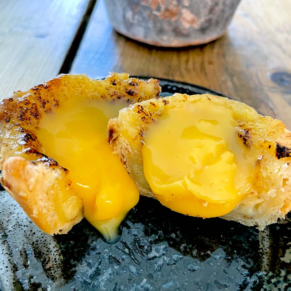A close-up of a pastry cut in half, revealing a rich, yellow, creamy filling oozing out onto a dark ceramic plate. The pastry has a golden, flaky crust and sits on a wooden table.