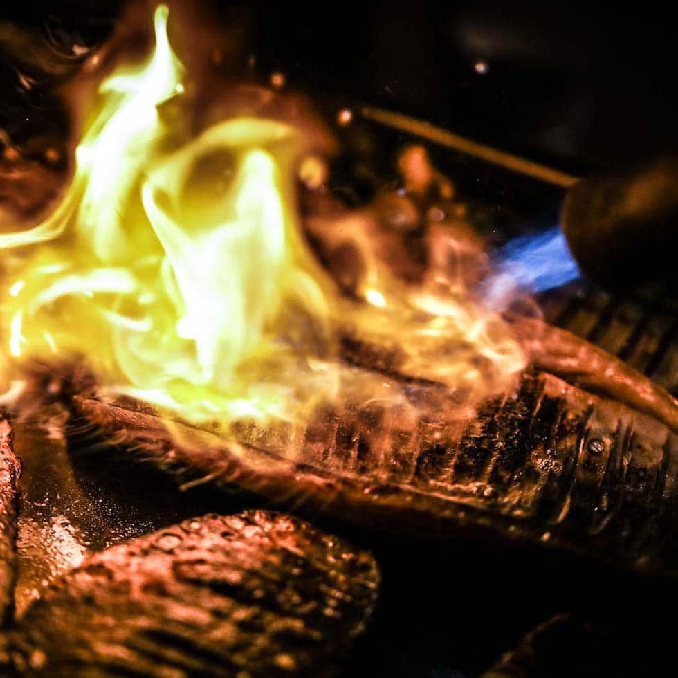 Slices of fish being seared with an open flame, creating a dramatic burst of yellow and orange fire over the surface, highlighting the texture and cooking process in a close-up shot.